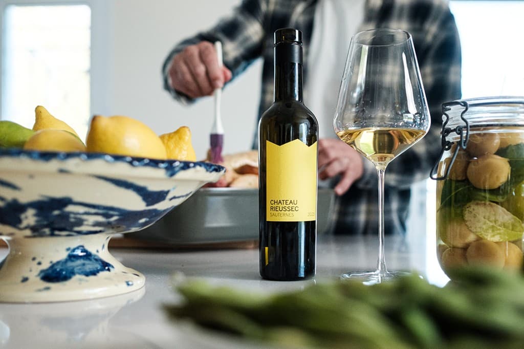 Bouteille de Château Rieussec 2024 Sauternes posée sur un plan de travail de cuisine, accompagnée d’un verre de vin doré, d’un compotier de citrons et d’un bocal de fruits confits